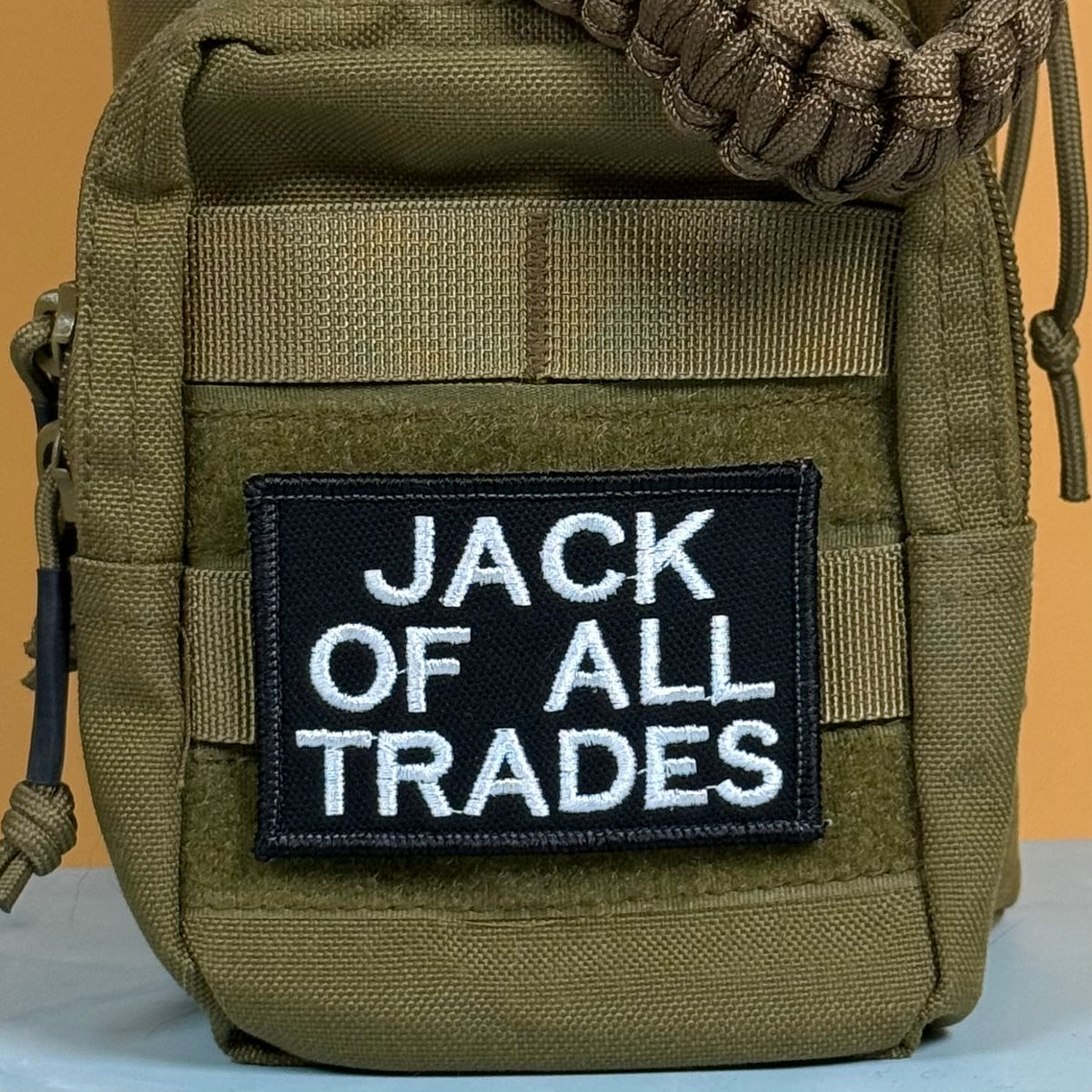 Jack Of All Trades embroidered morale patch: olive green tactical pouch with a black rectangular patch that reads 'JACK OF ALL TRADES' in white embroidered letters, shown against a plain orange and white background.