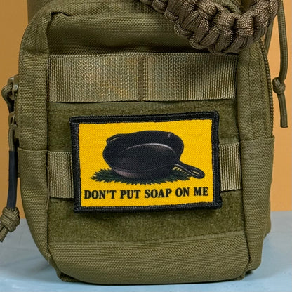 An olive green tactical pouch featuring a yellow rectangular patch with a black cast iron skillet and the text 'DON'T PUT SOAP ON ME'.