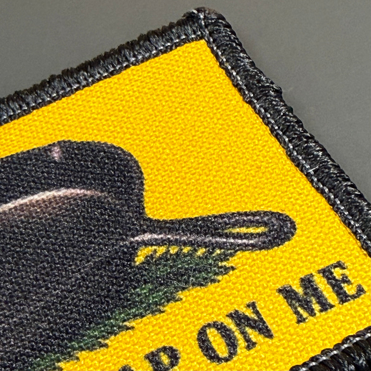 Close-up of a woven embroidered patch with a bright yellow field, black border, a black silhouette shape, green brushstroke detail, and partial black text reading "ON ME" on a gray background.