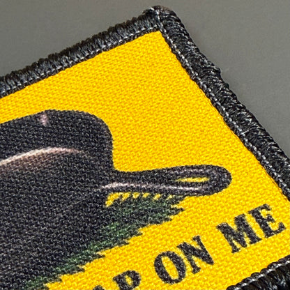 Close-up of a woven embroidered patch with a bright yellow field, black border, a black silhouette shape, green brushstroke detail, and partial black text reading "ON ME" on a gray background.