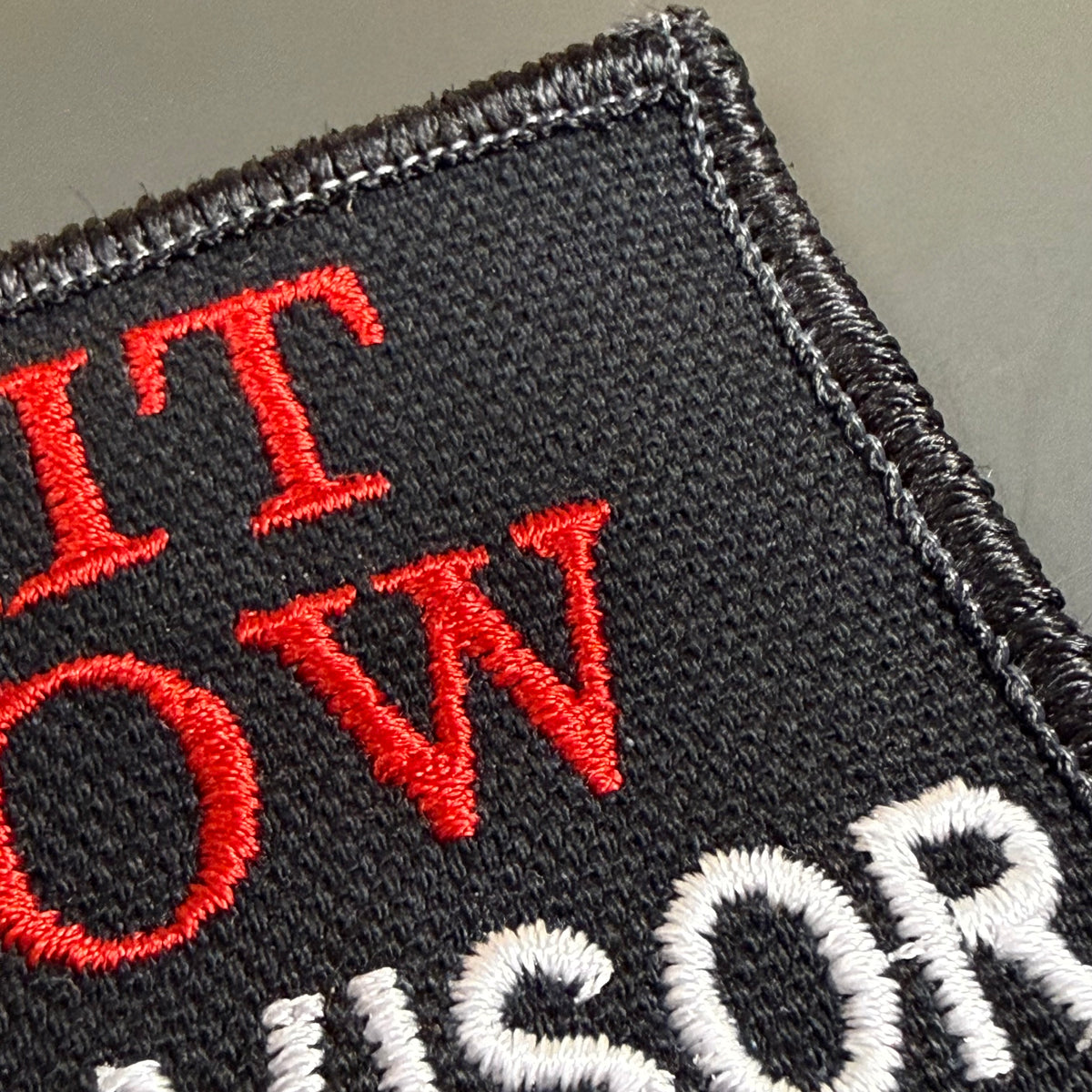 Close-up of an embroidered patch with black background, red lettering reading 'IT OW', and white lettering below, showing a stitched fabric border.