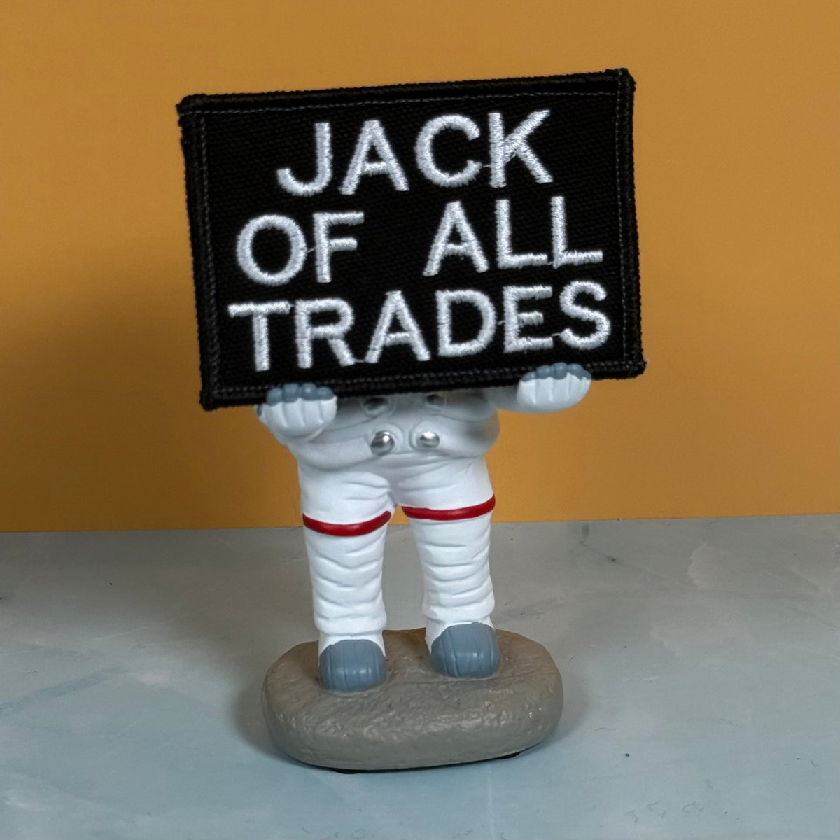 Jack Of All Trades embroidered morale patch: novelty figurine of an astronaut in a white spacesuit holding a black rectangular patch with white embroidered text reading 'JACK OF ALL TRADES'. The astronaut stands on a gray rock.