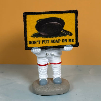 A small astronaut figurine holding a rectangular patch above its head. The patch is yellow with a black border, a black cast-iron skillet graphic, and the text 'DON'T PUT SOAP ON ME'.