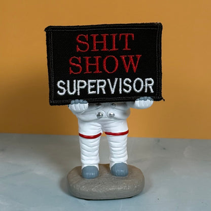 An embroidered patch reading 'SHIT SHOW SUPERVISOR' held by a small astronaut figurine on a gray base, set against a warm orange background.