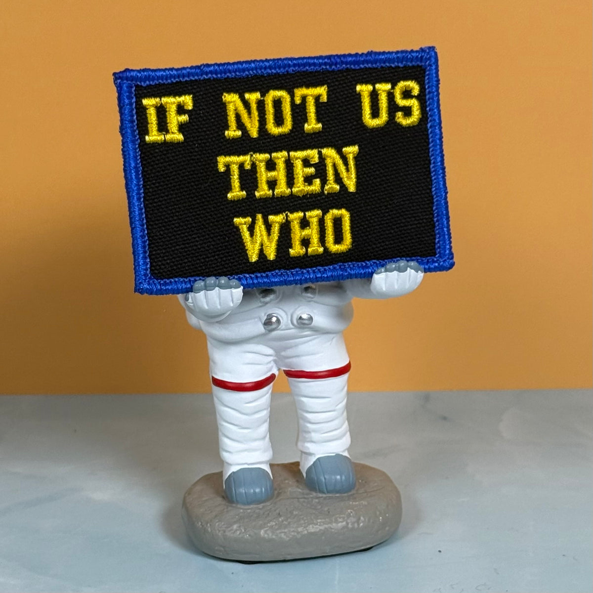 If Not Us Then Who embroidered morale patch: with the text 'IF NOT US THEN WHO' in yellow on a black background with a blue border, held by a small astronaut figurine on a gray base against a warm orange background.