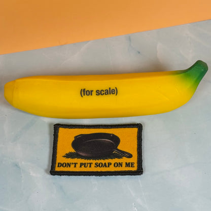 A yellow novelty squishy shaped like a banana with green tips, labeled '(for scale)', shown next to a rectangular embroidered patch that reads 'DON'T PUT SOAP ON ME' with a black frying pan graphic.