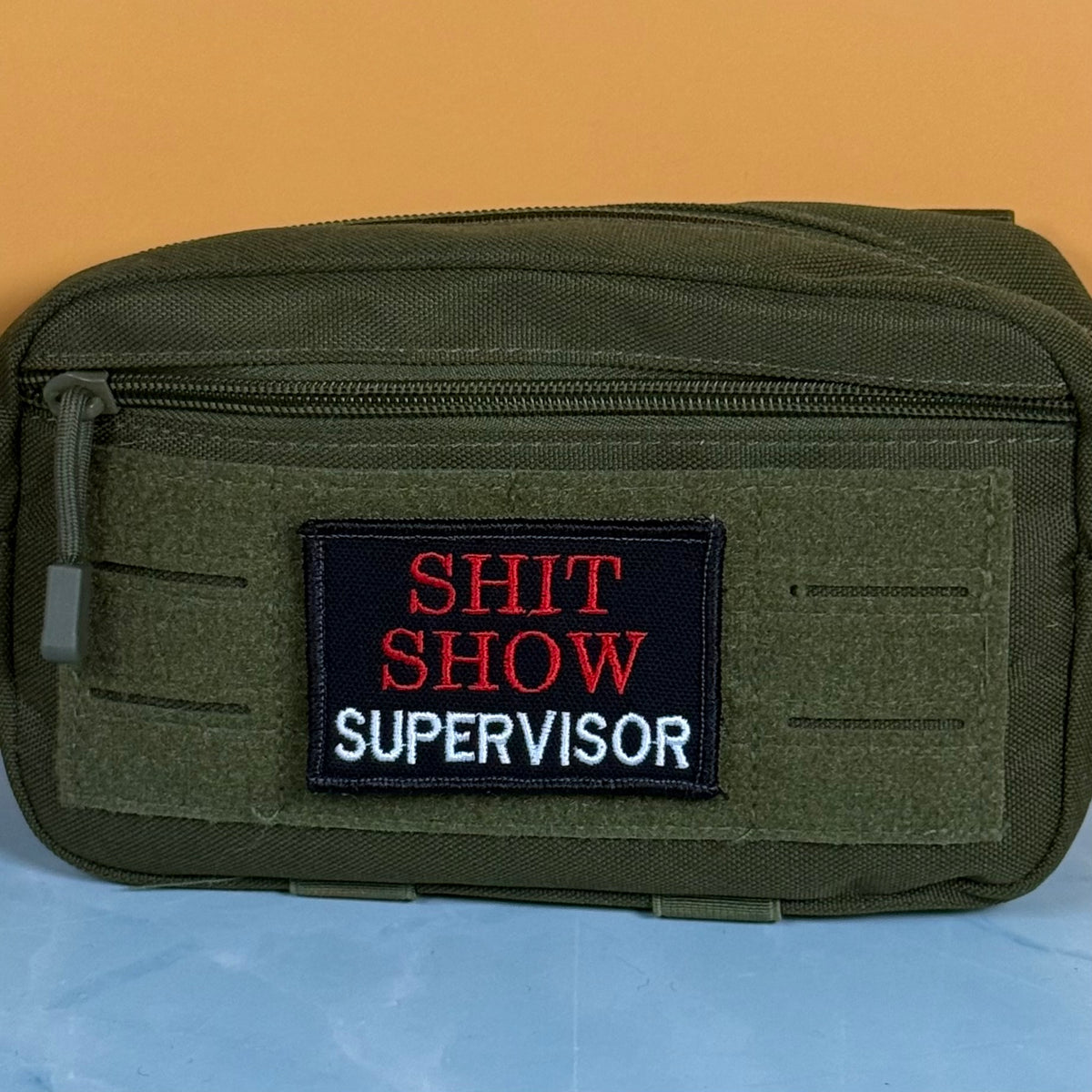 An olive green tactical pouch with a front zipper and a rectangular embroidered patch reading 'SHIT SHOW SUPERVISOR' in red and white on a black background, set against a solid orange backdrop.