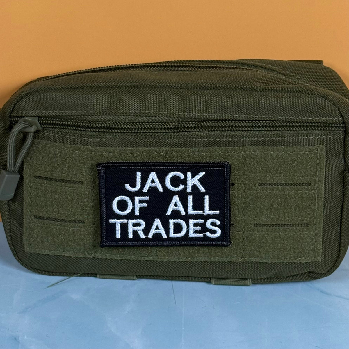 Jack Of All Trades embroidered morale patch: olive green tactical pouch with a black rectangular patch featuring the white embroidered text 'JACK OF ALL TRADES', set against a split orange and light blue background.