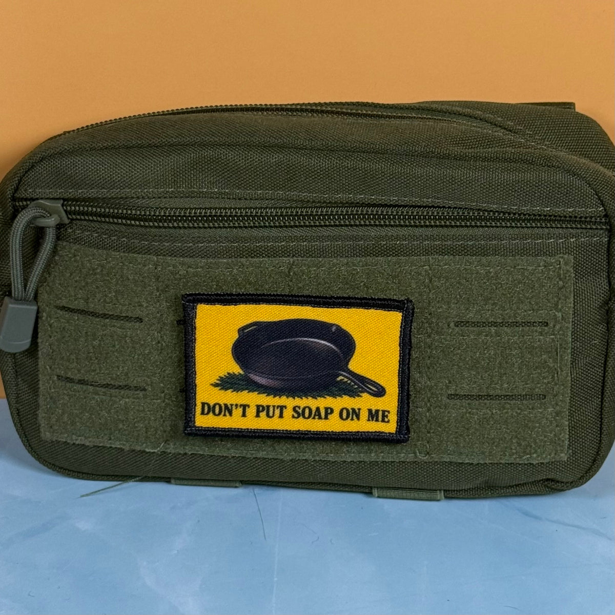An olive green tactical pouch with a yellow rectangular patch that reads 'DON'T PUT SOAP ON ME' and shows a black skillet graphic, set against a split orange and blue background.