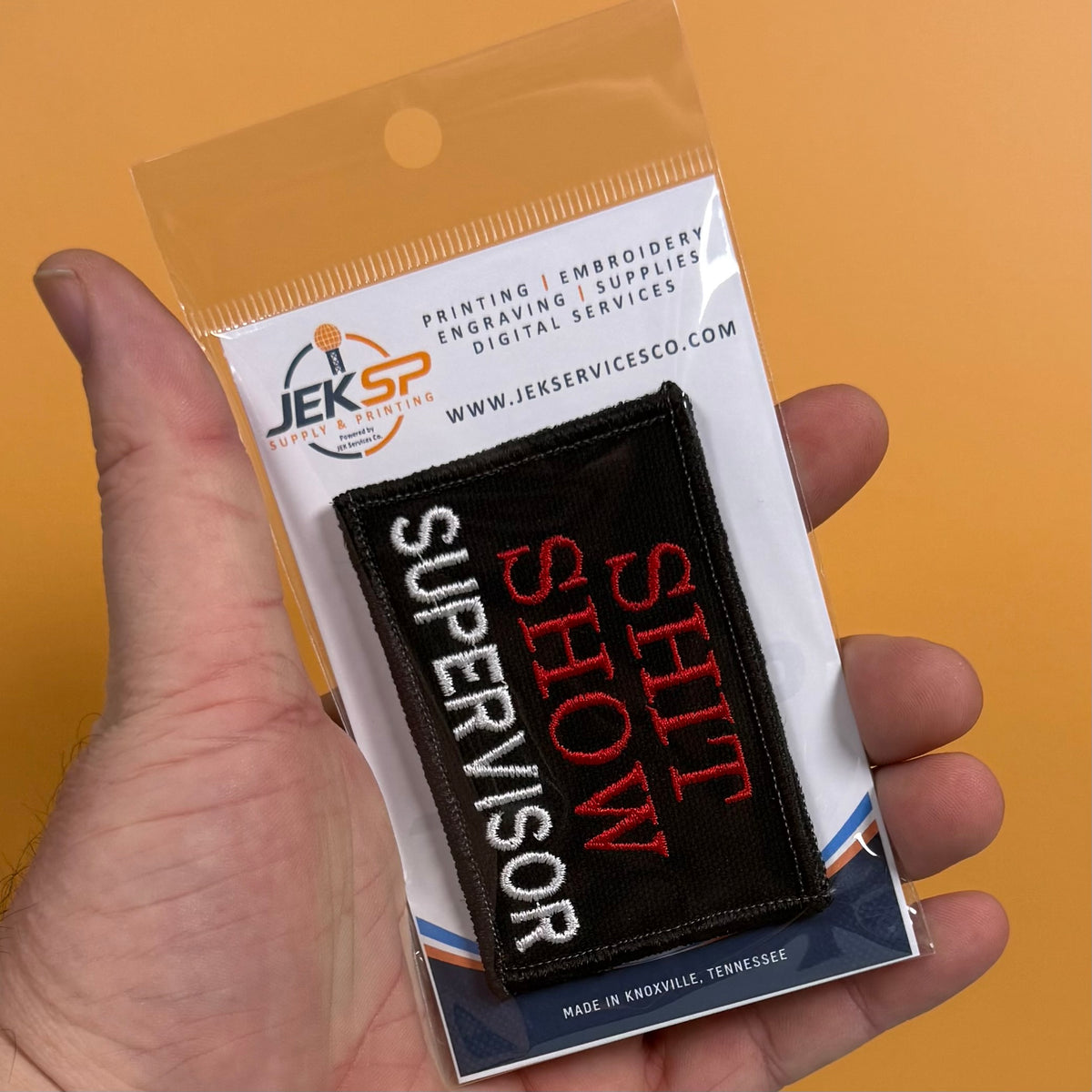 A hand holding a packaged embroidered patch that reads 'SUPERVISOR SHIT SHOW' in white and red on a black background, with the Jeksp Engraving Supplies logo visible on the card.