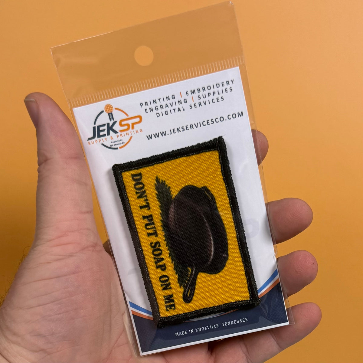 A hand holds a clear plastic blister pack containing an embroidered patch. The patch is yellow with a black border, a black pan graphic, and the text 'DON'T PUT SOAP ON ME' in black.