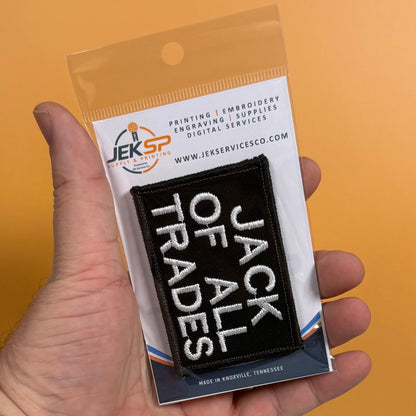 Jack Of All Trades embroidered morale patch: hand holds a packaged embroidered patch that reads “JACK OF ALL TRADES” in white text on a black background. The package card shows the JKS Services Co branding and product details.