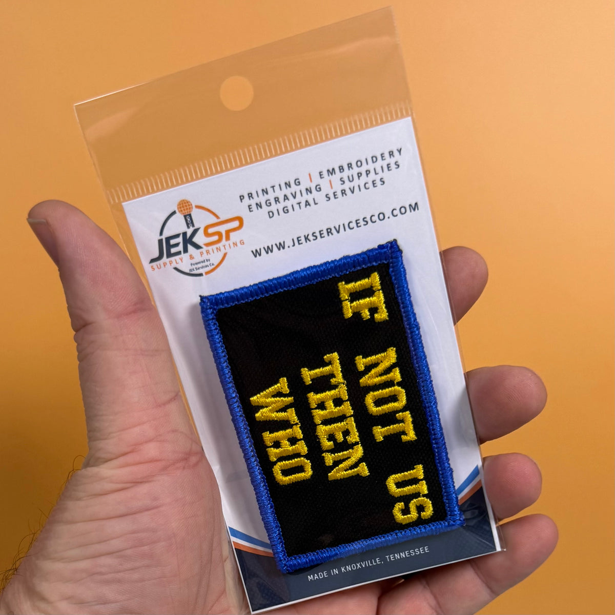 If Not Us Then Who embroidered morale patch: hand holds a packaged embroidered patch against a solid orange background. The patch is black with yellow text reading 'IF NOT US THEN WHO' and a blue border.