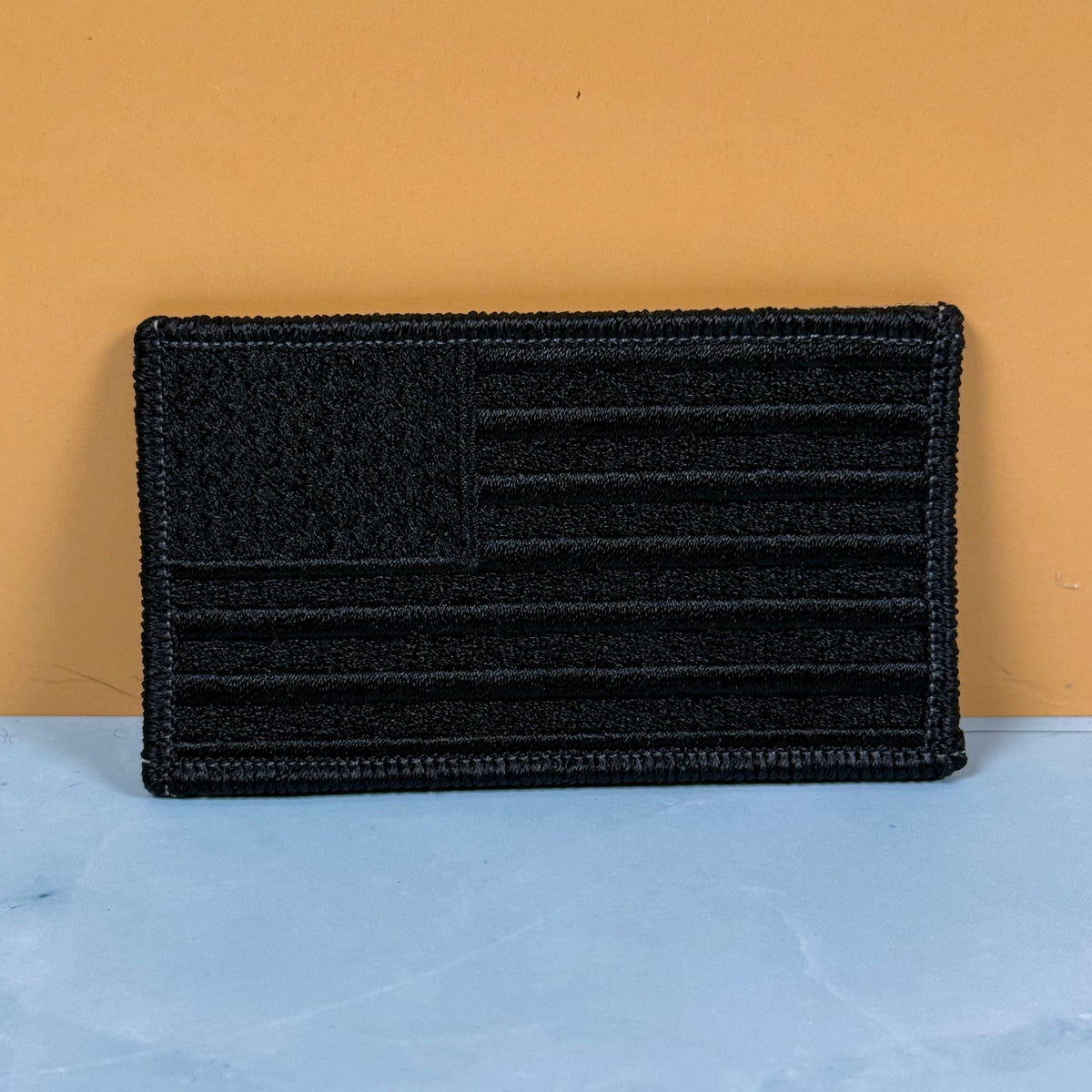 Blackout No Quarters Given American Flag embroidered morale patch: embroidered American flag patch with a hook-and-loop style design, displayed against a split orange and light blue background.