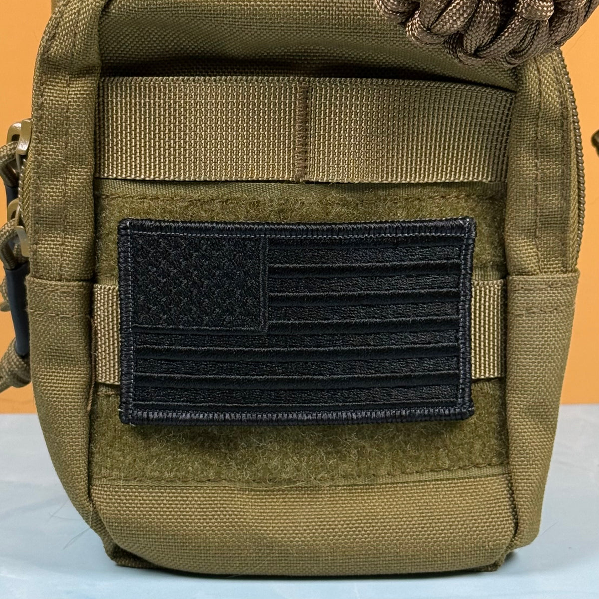 Blackout No Quarters Given American Flag embroidered morale patch: tactical pouch in olive drab green with a black embroidered American flag patch on the front, set against a solid orange background.
