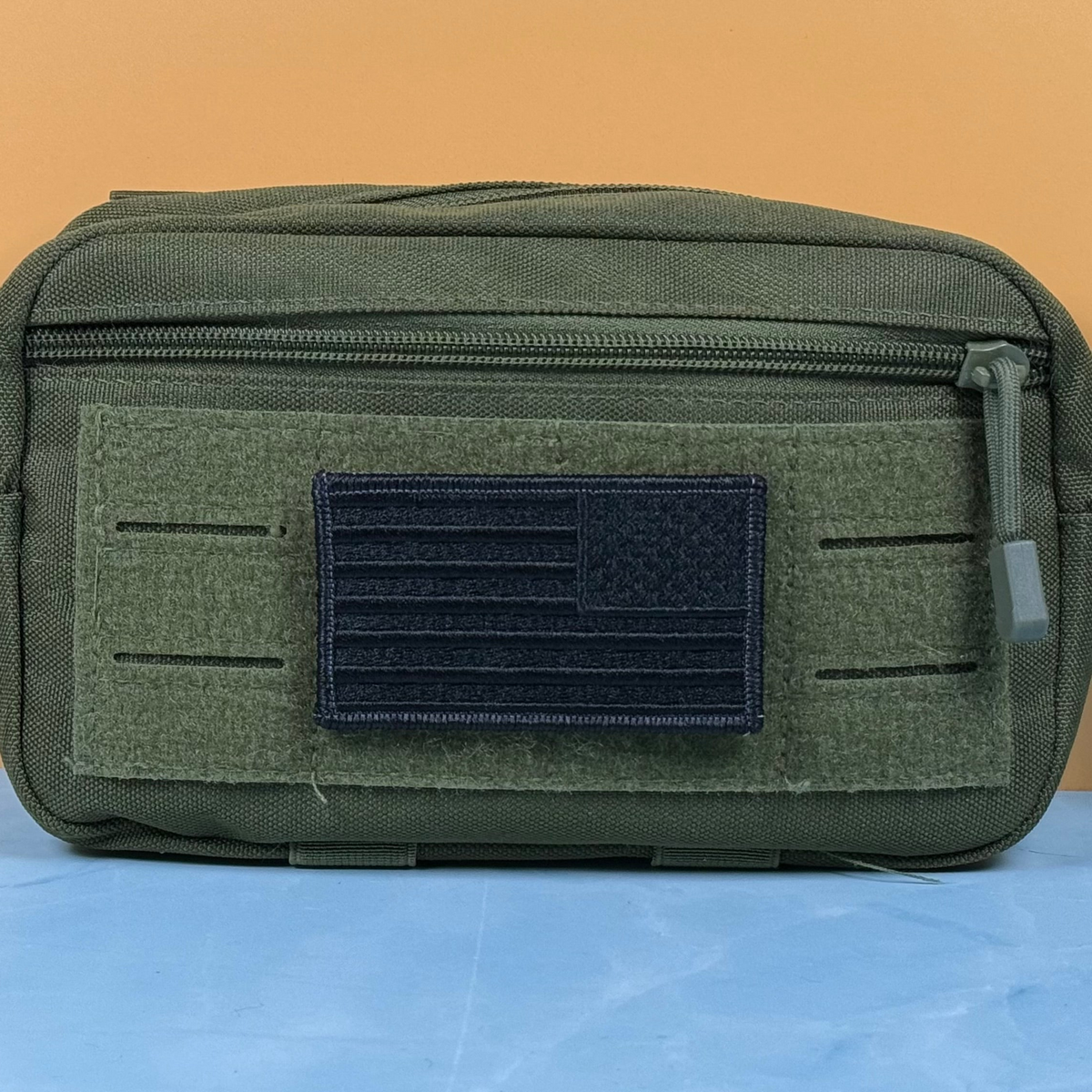 Blackout No Quarters Given American Flag embroidered morale patch: olive green tactical pouch with a front zippered pocket and a dark navy American flag patch on a hook-and-loop panel, set against a split orange and blue background.