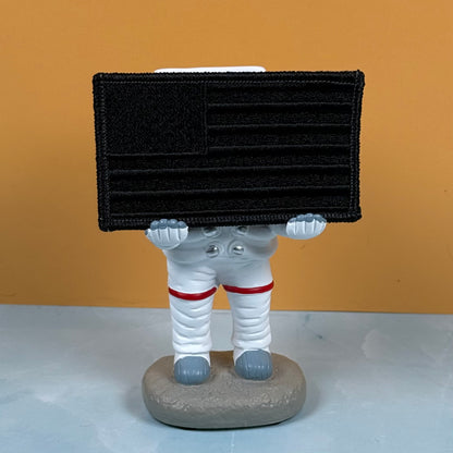 Blackout No Quarters Given American Flag embroidered morale patch: astronaut figurine holding a black rectangular patch with an American flag design, standing on a gray base against a warm orange background.