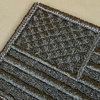 Blackout No Quarters Given American Flag embroidered morale patch: dark embroidered patch with a textured flag design, featuring a black and gray American flag pattern on a beige background.