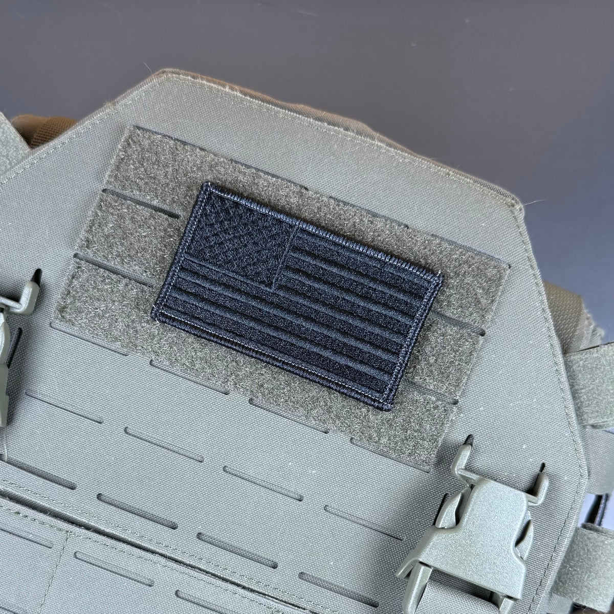Blackout No Quarters Given American Flag embroidered morale patch: dark gray embroidered American flag patch mounted on a gray tactical vest with hook-and-loop fastening, shown in a close-up product detail shot.