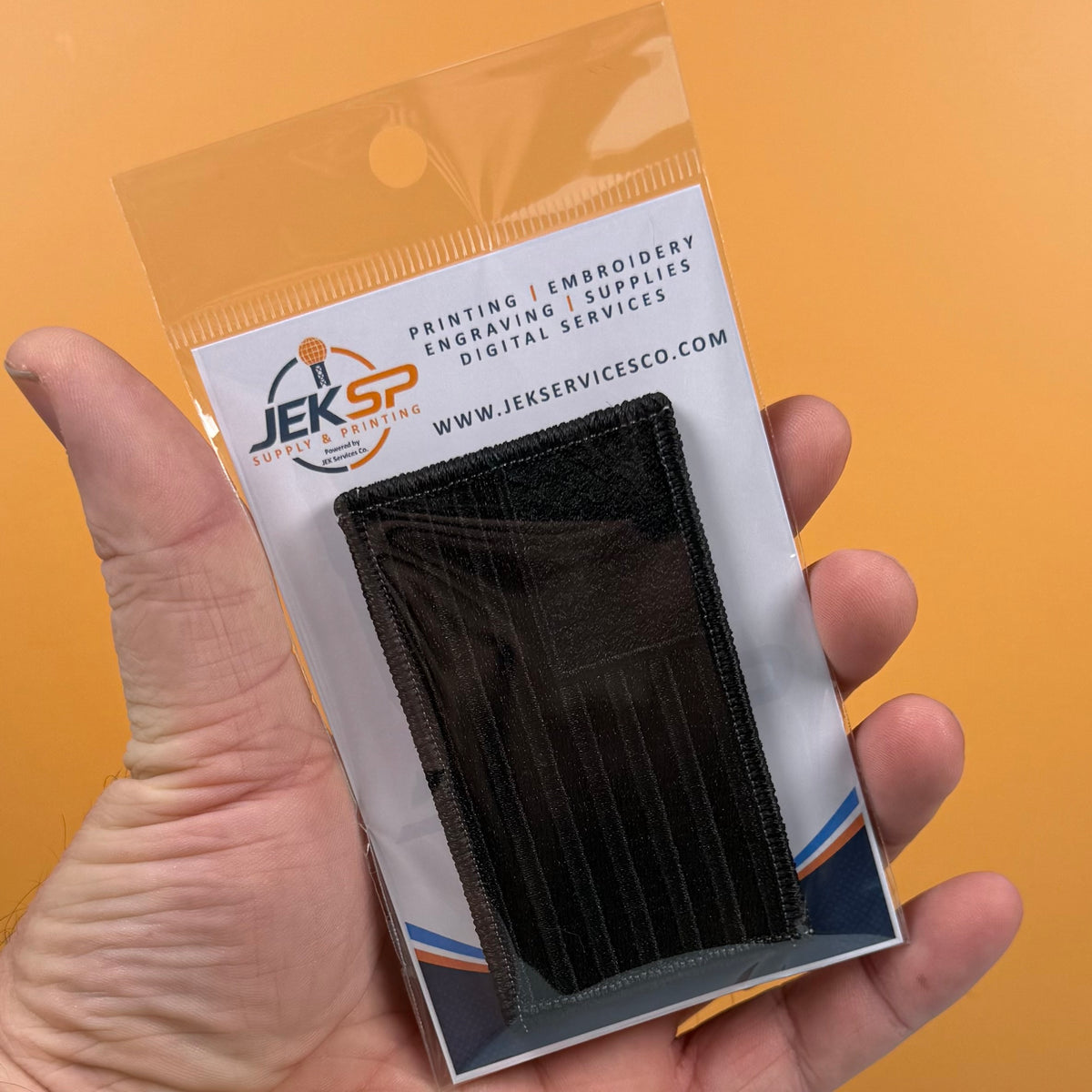 Blackout No Quarters Given American Flag embroidered morale patch: hand holding a small black embroidered pouch inside a clear plastic retail package with a white backing card. The card shows the JEKSP Digital Services logo and website,.