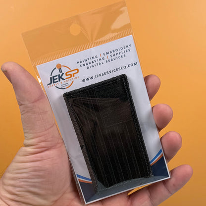 Blackout No Quarters Given American Flag embroidered morale patch: hand holding a small black embroidered pouch inside a clear plastic retail package with a white backing card. The card shows the JEKSP Digital Services logo and website,.