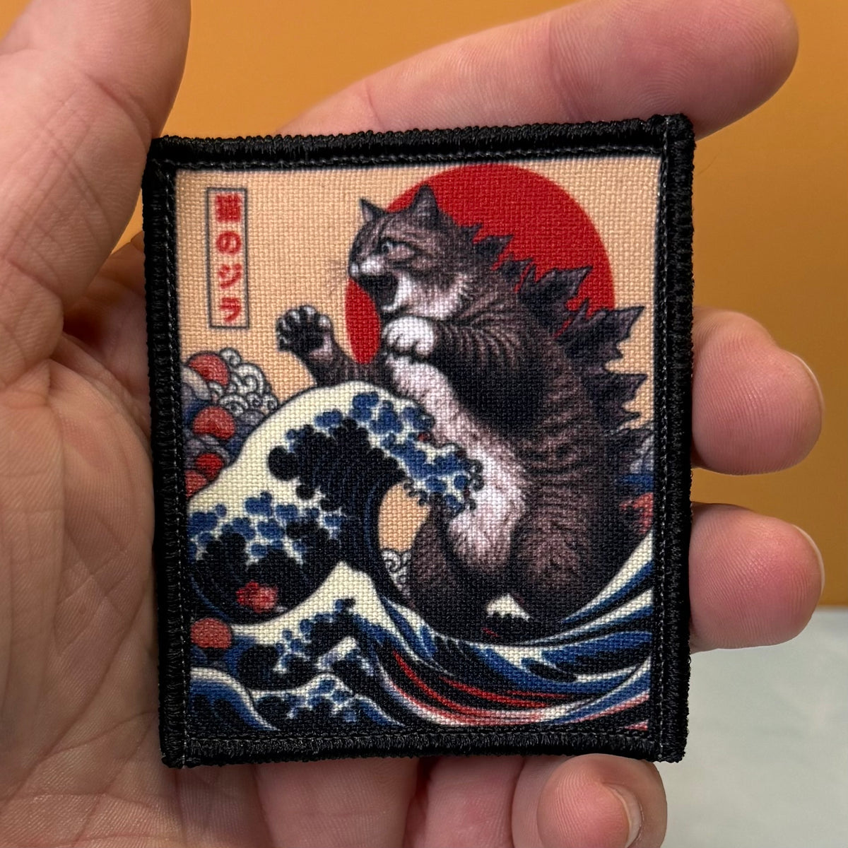 Catzilla printed canvas morale patch held in a hand, featuring a cat in the style of Hokusai's Great Wave off Kanagawa with a red sun and Japanese text.