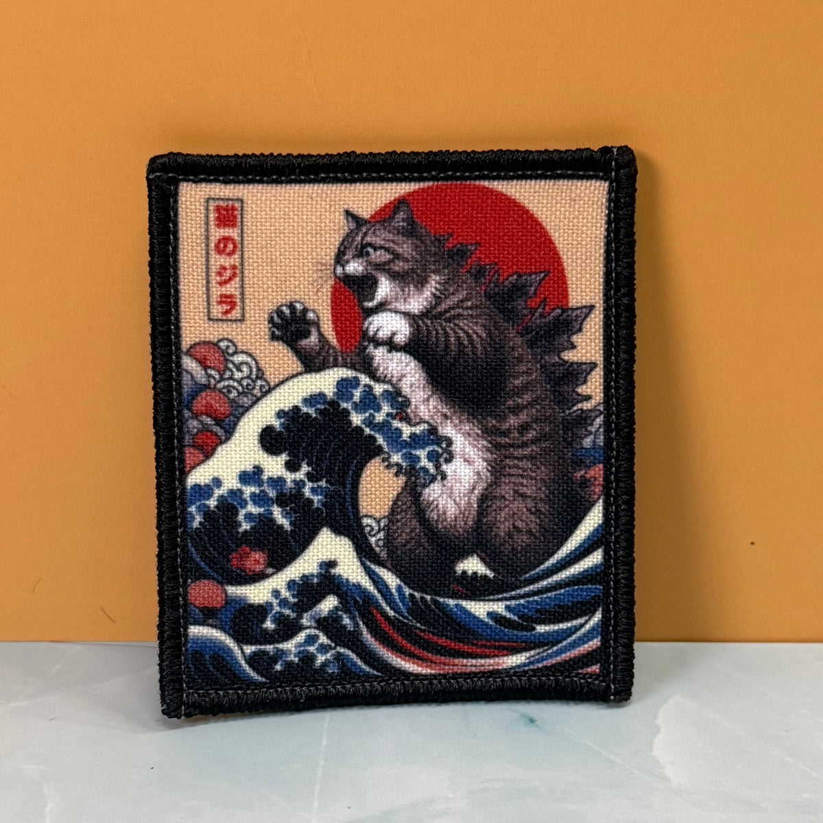Catzilla printed canvas morale patch featuring a tabby cat in a Godzilla-like pose rising from blue and white waves, with a large red sun and Japanese text on a beige background, bordered by black stitching. The patch is.