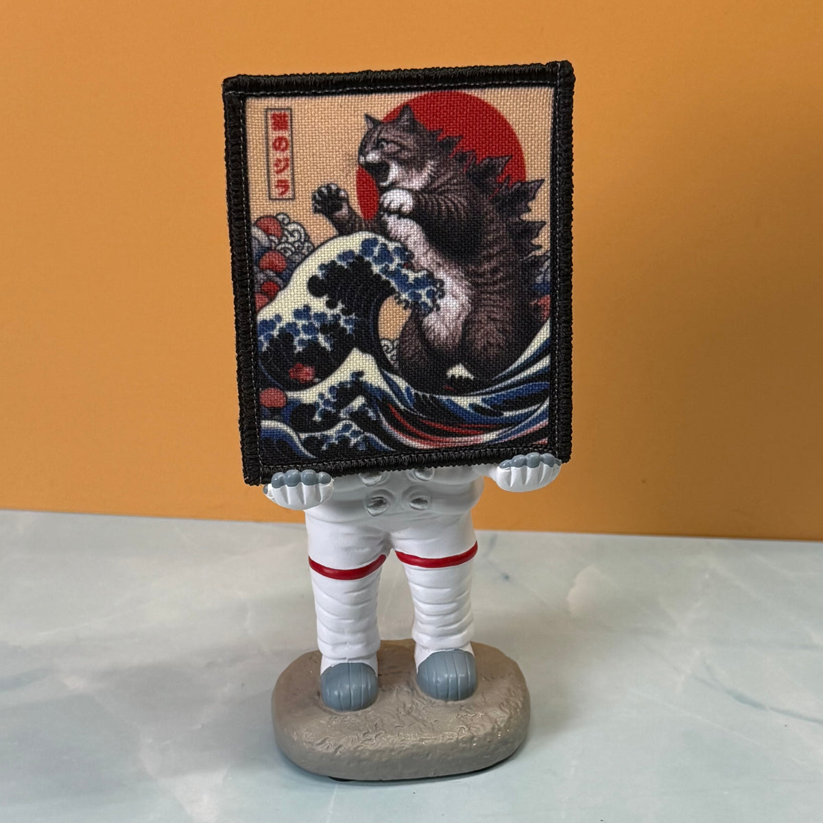 Catzilla printed canvas morale patch, a small astronaut figurine standing on a round base, holding a square printed patch or tapestry in front of its face. The patch shows a cat riding a large blue wave with a red sun and.