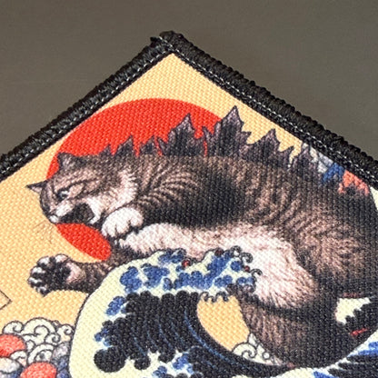 Catzilla printed canvas morale patch, a close-up of a fabric patch featuring a tabby cat in a dynamic pose over a blue and white wave, with a red sun and dark mountain silhouette on a beige background, finished with black.