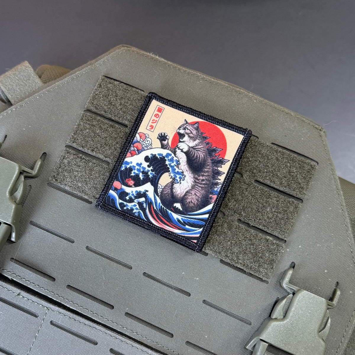Catzilla printed canvas morale patch featuring a Japanese ukiyo-e style cat under a red sun and blue waves, mounted on olive drab MOLLE webbing of a tactical vest.