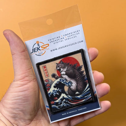 Catzilla printed canvas morale patch, a hand holds a packaged printed patch featuring a cat riding a wave in a Japanese ukiyo-e style, with a JEKSP logo on the backing card against an orange background.