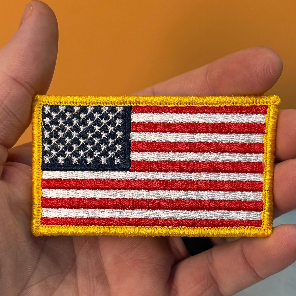 American Flag embroidered morale patch: United States flag patch with a yellow border, held in a person's hand against a solid orange background.