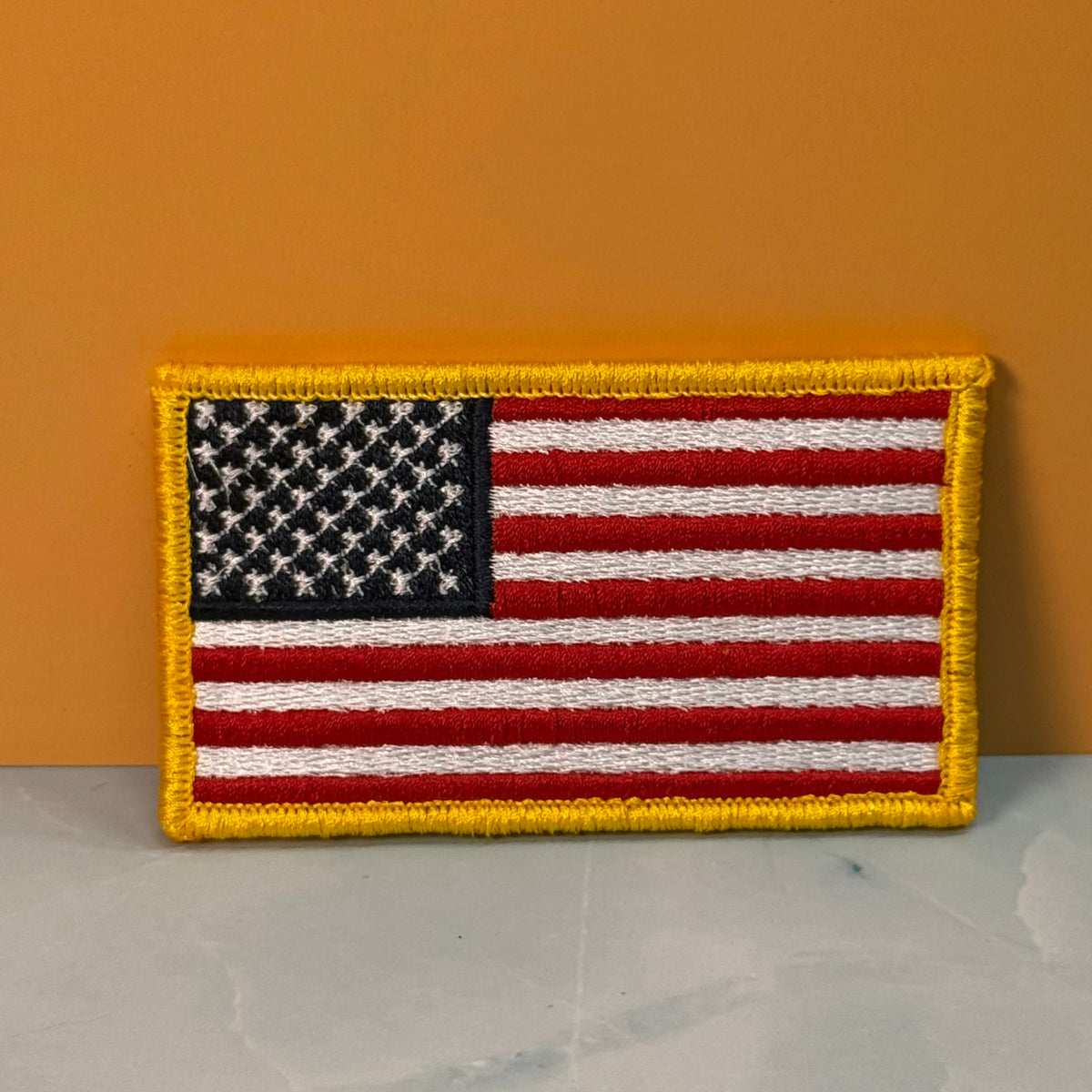 American Flag embroidered morale patch: United States flag patch with a yellow border, displayed against a solid orange background and a light gray marble surface.