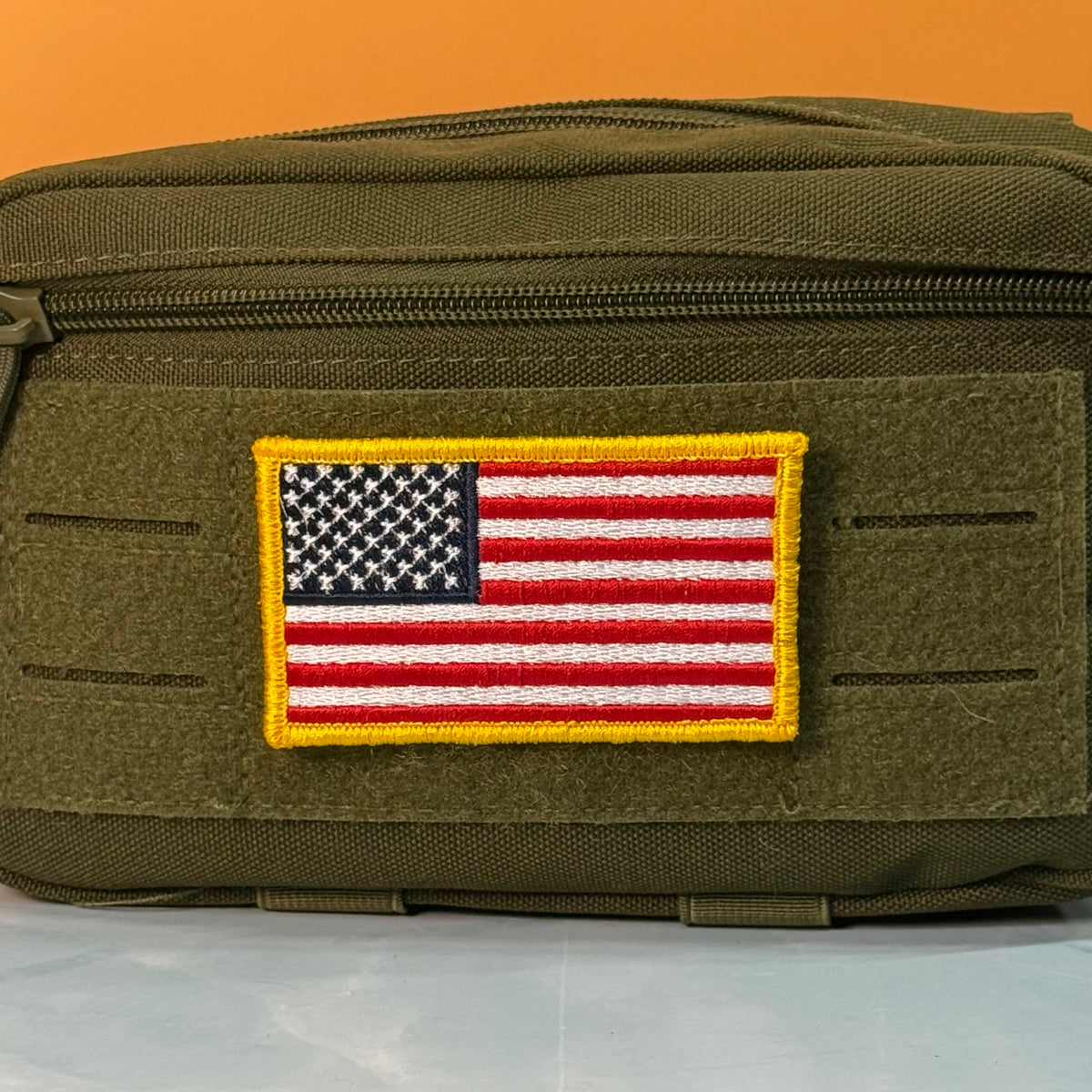 American Flag embroidered morale patch: olive drab tactical pouch with a rectangular embroidered American flag patch on the front, set against a solid orange background.