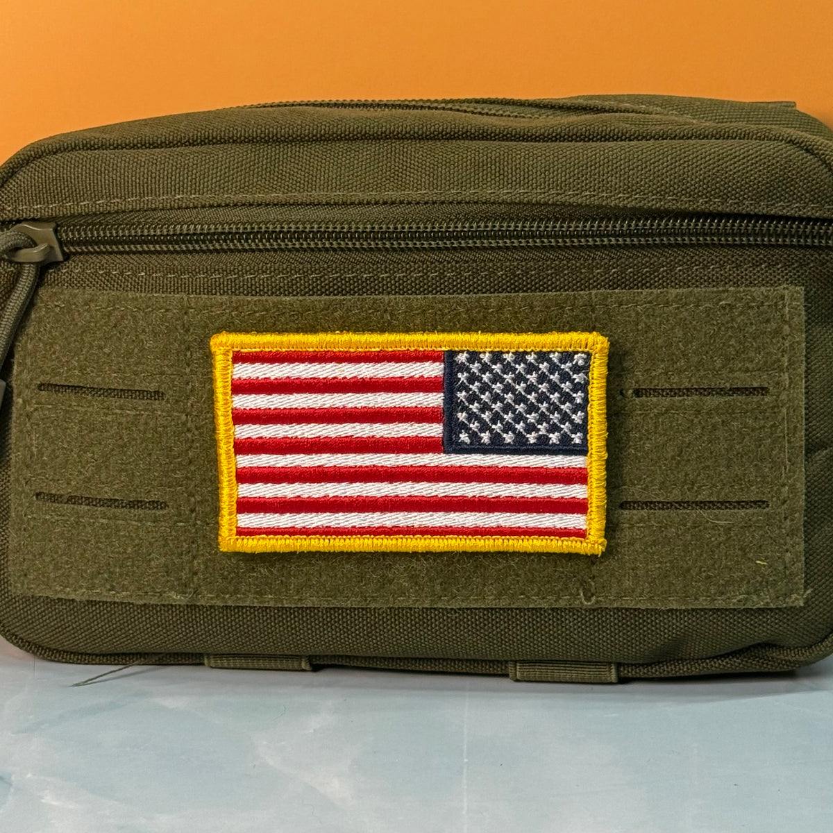 American Flag embroidered morale patch: olive green tactical pouch with a rectangular embroidered American flag patch on the front, set against a solid orange background.