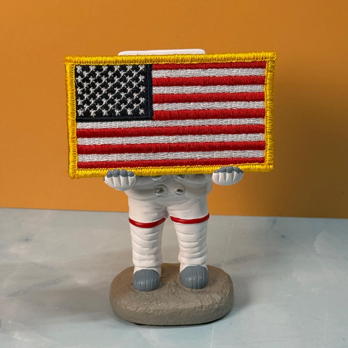 American Flag embroidered morale patch: astronaut figurine holding a large embroidered American flag patch over its head, standing on a gray moon-like base against an orange background.