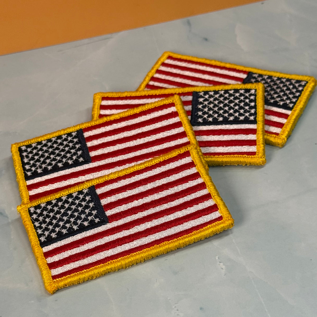 American Flag embroidered morale patch: Three embroidered American flag patches with gold borders, arranged diagonally on a light gray marble-like surface with a warm orange background visible in the top corner.