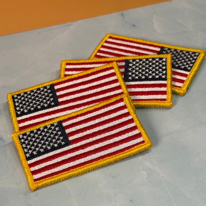 American Flag embroidered morale patch: Three embroidered American flag patches with gold borders, arranged diagonally on a light gray marble-like surface with a warm orange background visible in the top corner.