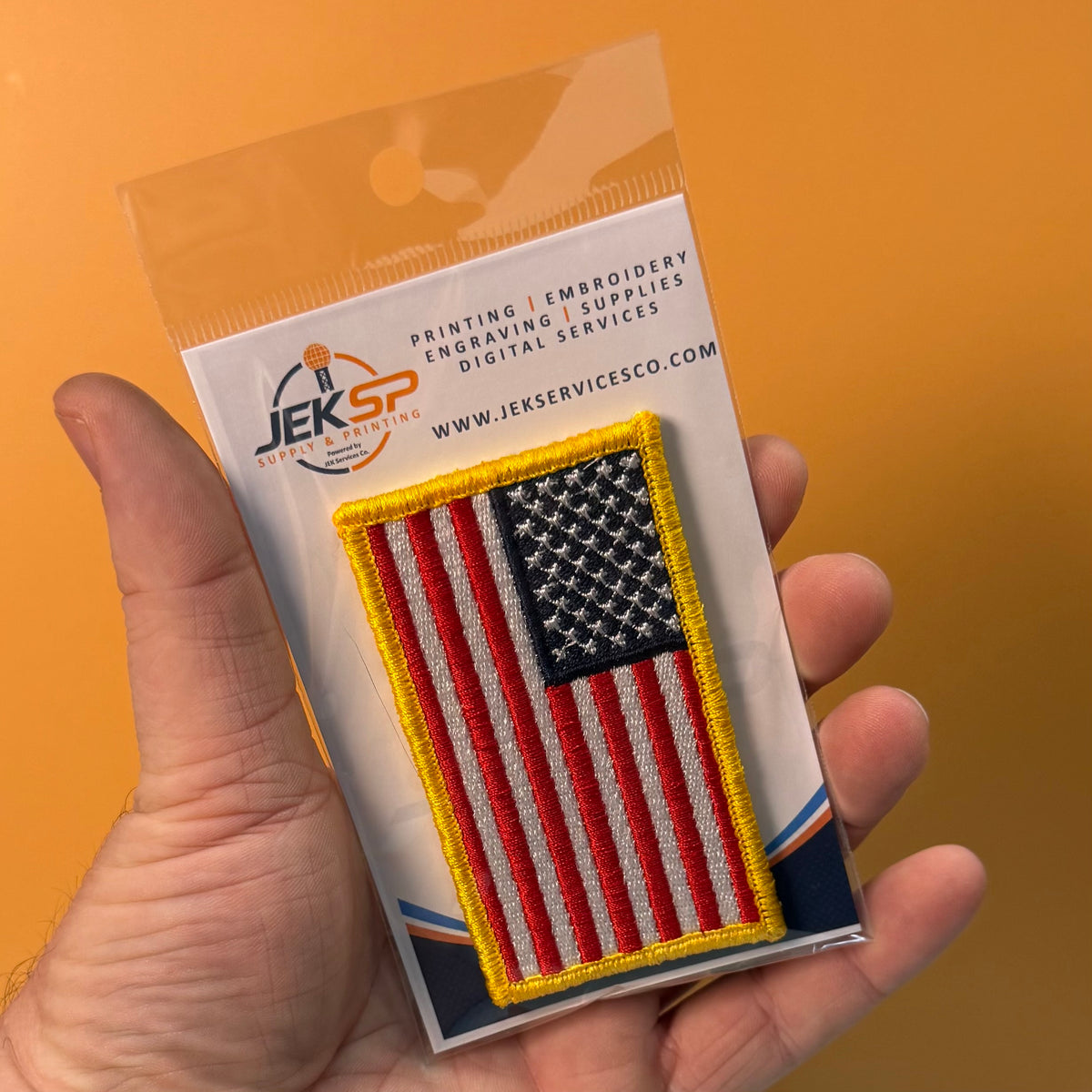 American Flag embroidered morale patch: hand holds a packaged embroidered American flag patch with red and white stripes, a blue star field, and a yellow border. The backing card shows the JEKSP logo and text for printing, embroidery,.