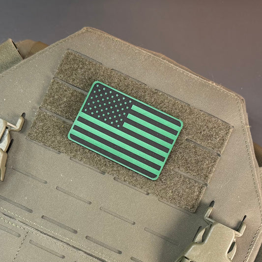 A rectangular tactical patch featuring a stylized American flag design in black and bright green, mounted on a tan tactical vest with hook-and-loop fastening.