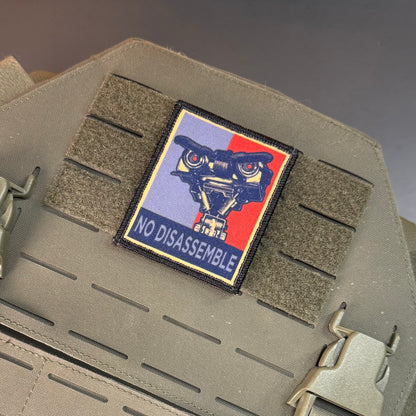 A rectangular tactical patch with a black border and the words 'NO DISASSEMBLE' is attached to olive drab webbing on a tactical vest. The patch features a stylized machine gun graphic split across light blue and red sections with a black background.