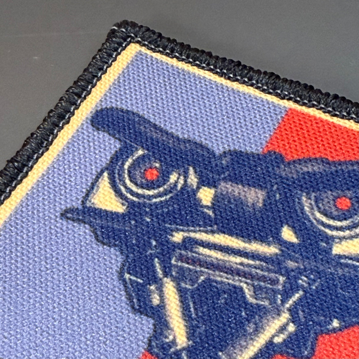 Close-up of a rectangular embroidered patch with a stylized robotic face. The patch has a black stitched border, a light blue field, red and yellow geometric sections, and a dark blue robot graphic with small red eyes.