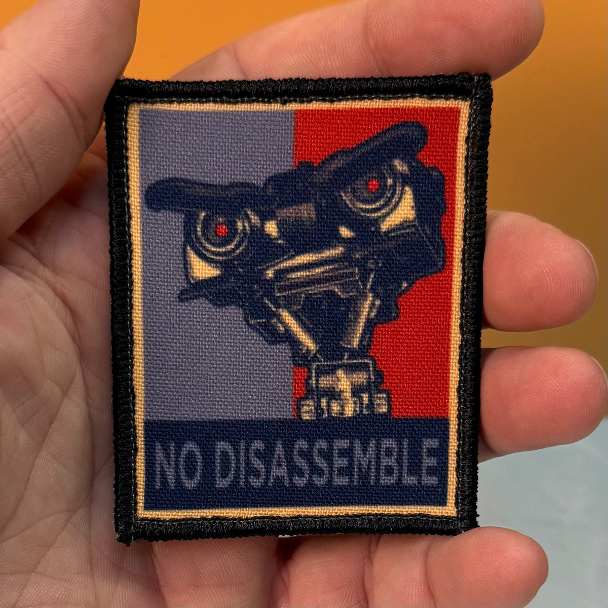 A hand holds a rectangular embroidered patch featuring a stylized robot face with red eyes and the text 'NO DISASSEMBLE' at the bottom. The patch has a black border and a split background in gray and red with a thin gold line.