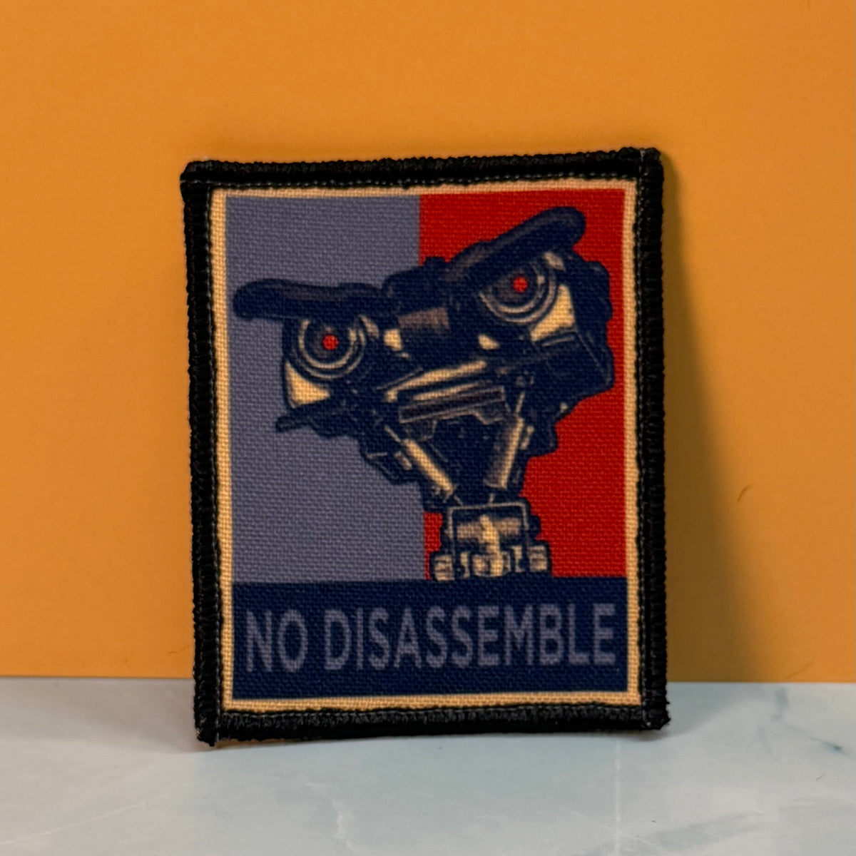 A square embroidered patch featuring a stylized robot face with red eyes on a split blue-gray and red background, with the text 'NO DISASSEMBLE' at the bottom, photographed against an orange wall and light marble surface.