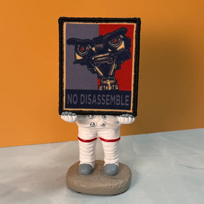 A small astronaut figurine standing on a round base, holding a square patch above its head. The patch has a black border and shows a robot face with the words 'NO DISASSEMBLE' across the bottom.