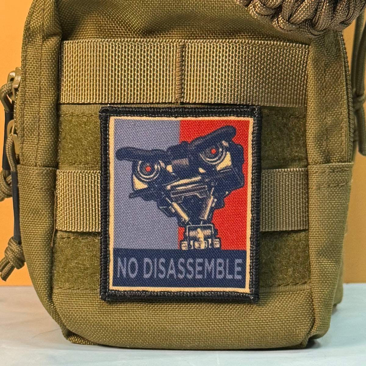 An olive drab tactical pouch featuring a rectangular embroidered patch with a robot face and the text 'NO DISASSEMBLE' on a plain background.