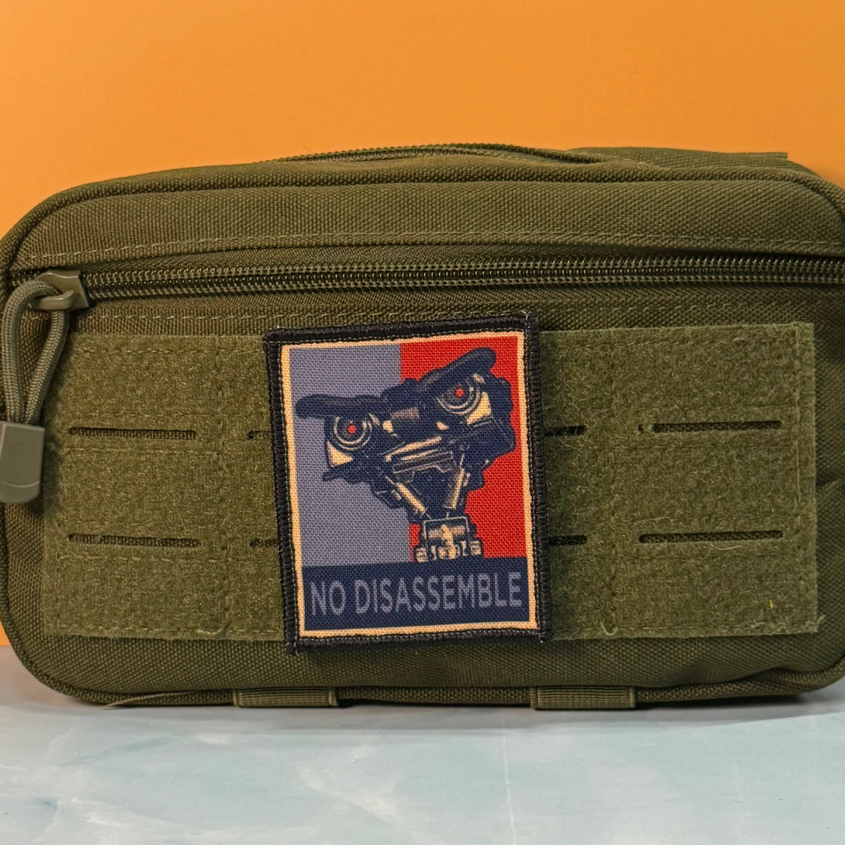 An olive green tactical pouch with a rectangular embroidered patch featuring a robot eye and the text 'NO DISASSEMBLE' on a blue and red background, set against an orange backdrop.