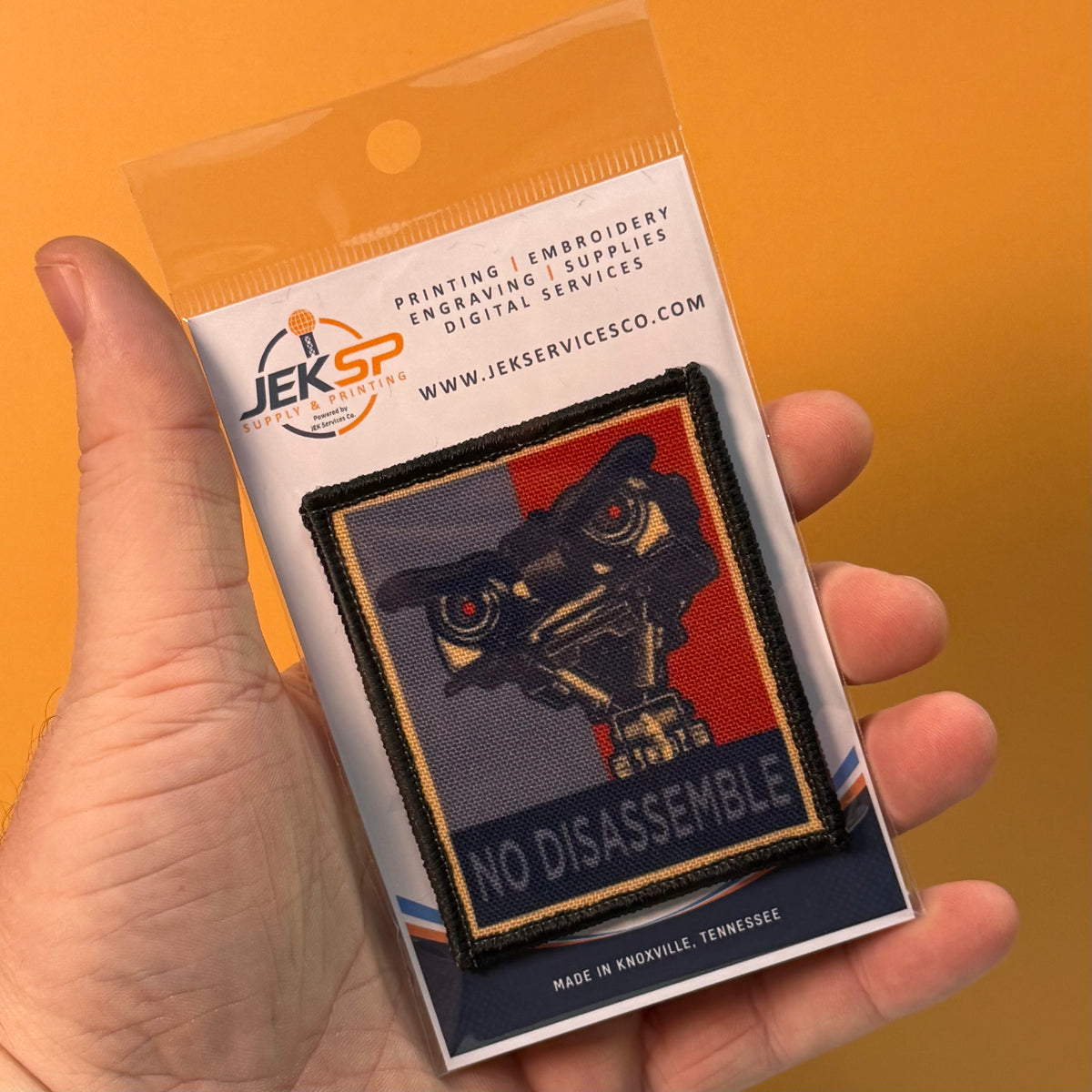 A hand holds a packaged embroidered patch against a solid orange background. The patch features a stylized robot head with red eyes and the words 'NO DISASSEMBLE' on a dark blue, red, and black design.