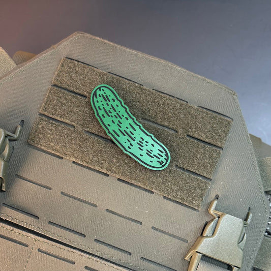 A green pickle-shaped patch with black dotted details, displayed on a gray tactical vest with a hook-and-loop panel.