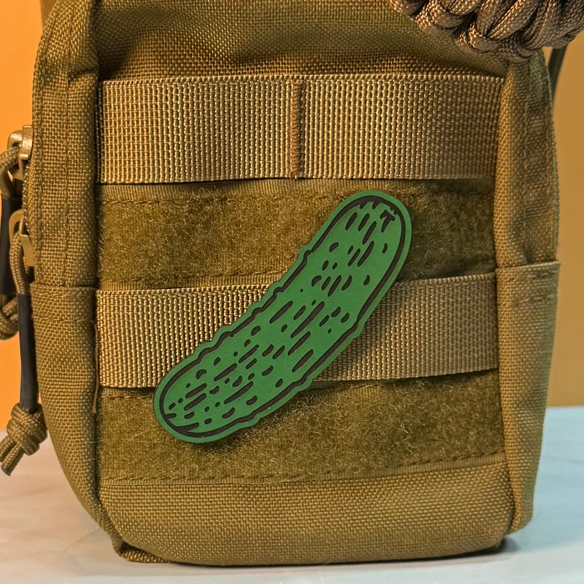 Dill Pickle laser-engraved synthetic morale patch: a green laser-engraved synthetic patch shaped like a cucumber or pickle with black textured lines, shown attached to the side webbing of a tan tactical bag.